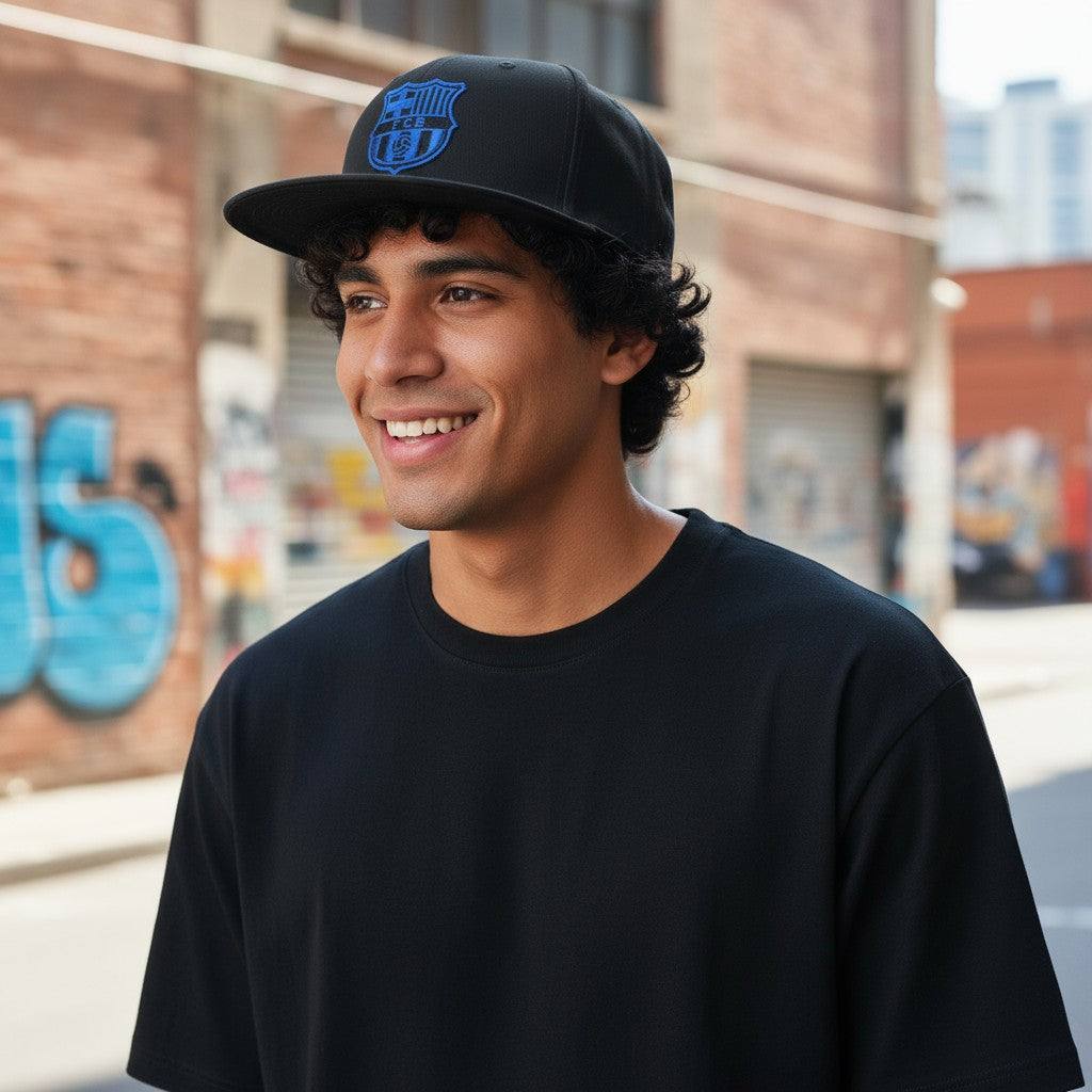 Young man wearing a black Barcelona Cap Locale Snapback Hat with blue logo and flat brim outdoors in urban setting